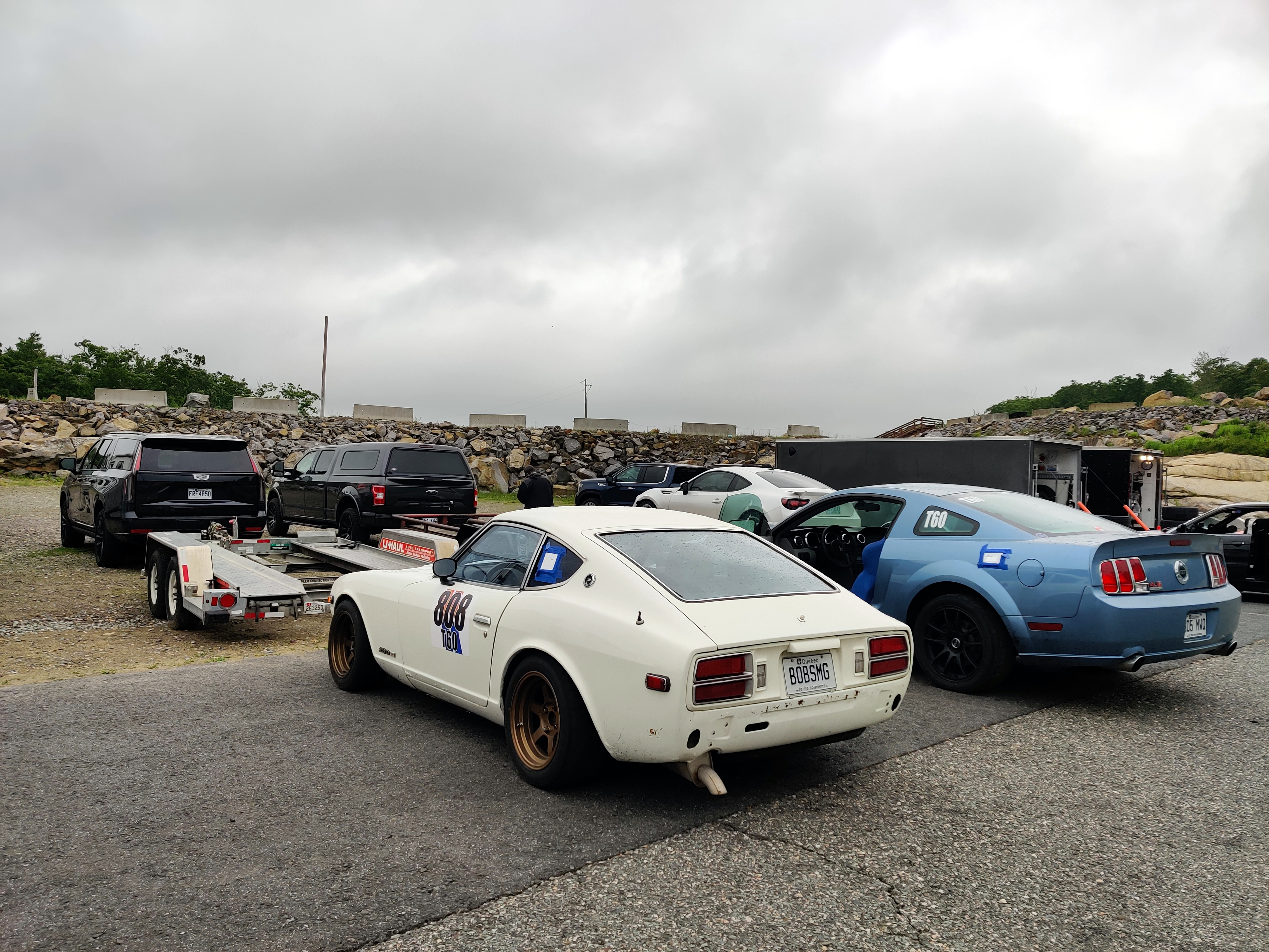 A 1978 Datsun 280Z rests on pavement behind a trailer and a black 2023 Cadillac Escalade
