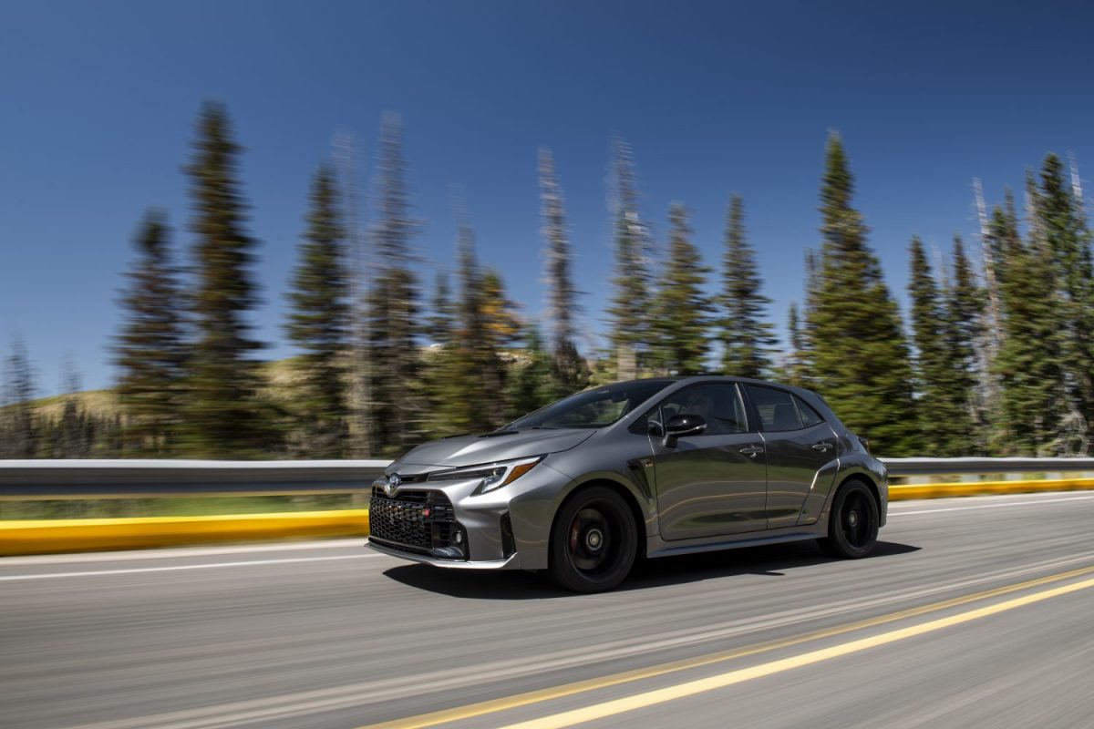 A 2023 Toyota GR Corolla in "heavy metal" gray chrome paint speeds across a highway past a collection of tall trees