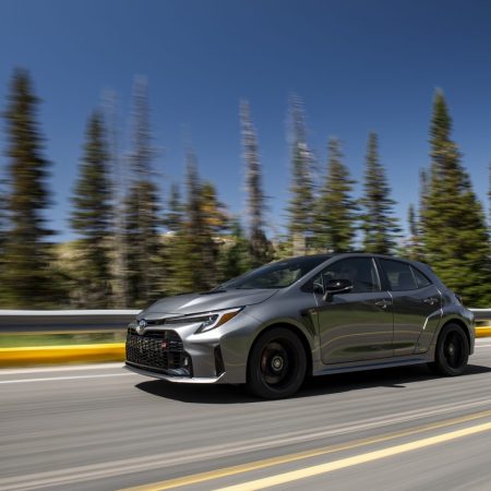 A 2023 Toyota GR Corolla in "heavy metal" gray chrome paint speeds across a highway past a collection of tall trees