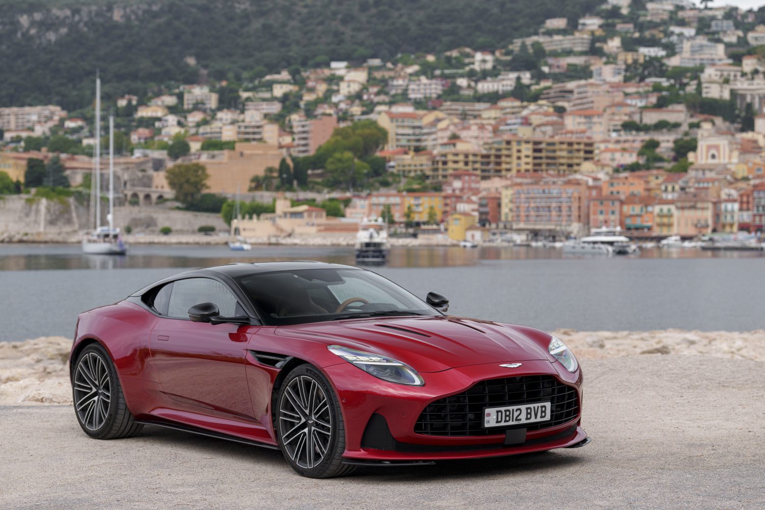 The Aston Martin DB12 in hyper red, parked across the river of a coastline village
