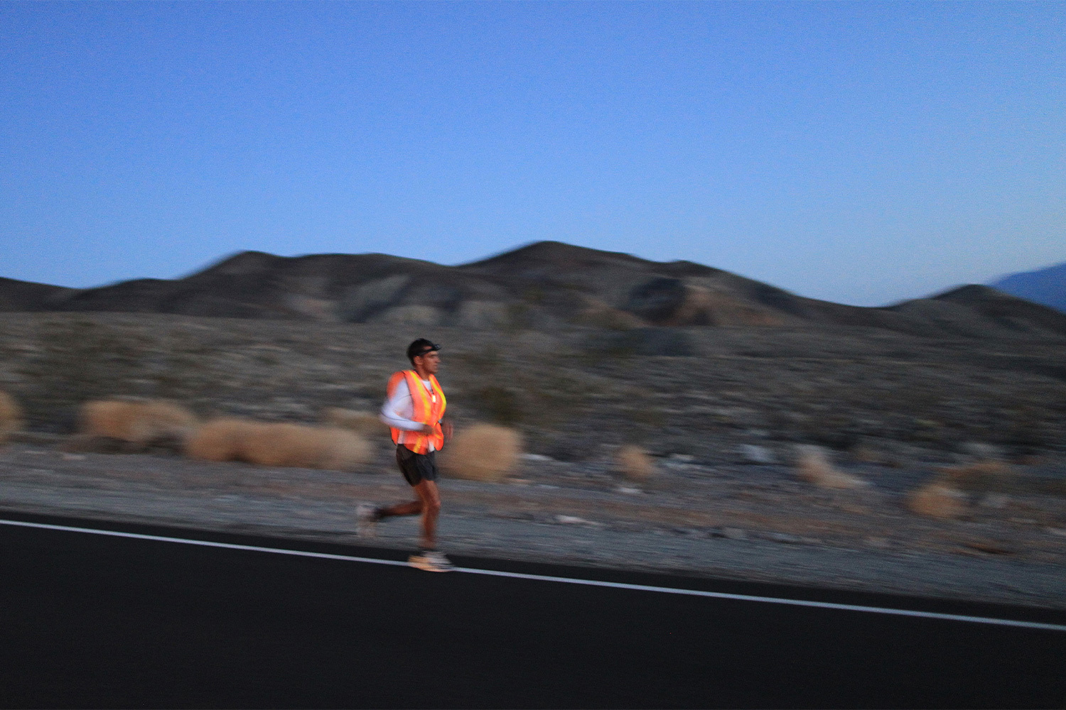 A man running through the desert at dusk in a neon orange singlet during an endurance race.