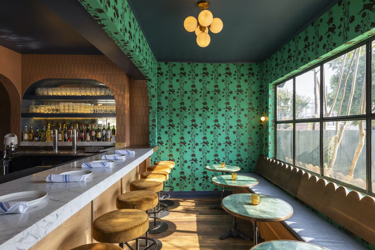 Seating area with booth seats at a window, a bar with barstools and green wallpaper