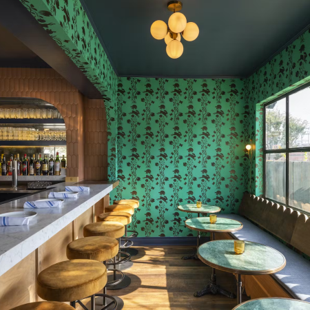 Seating area with booth seats at a window, a bar with barstools and green wallpaper