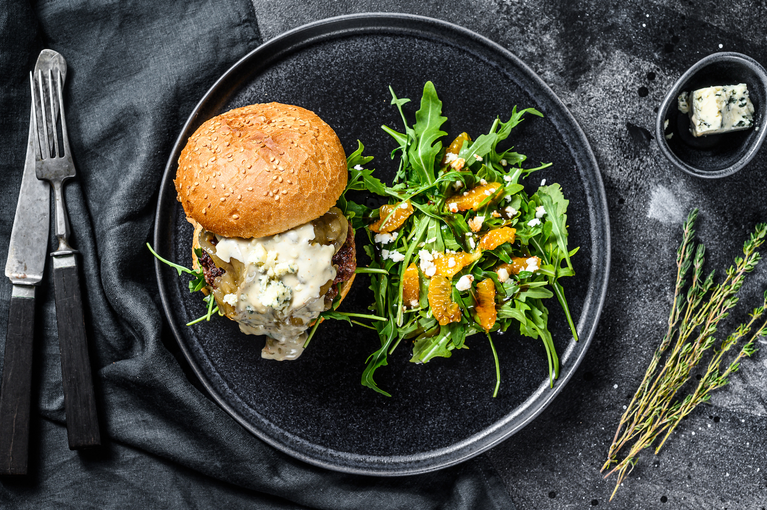 Burgers with blue cheese, bacon, marbled beef and onion marmalade, a side dish of salad with arugula and oranges. Black background. Top view 
