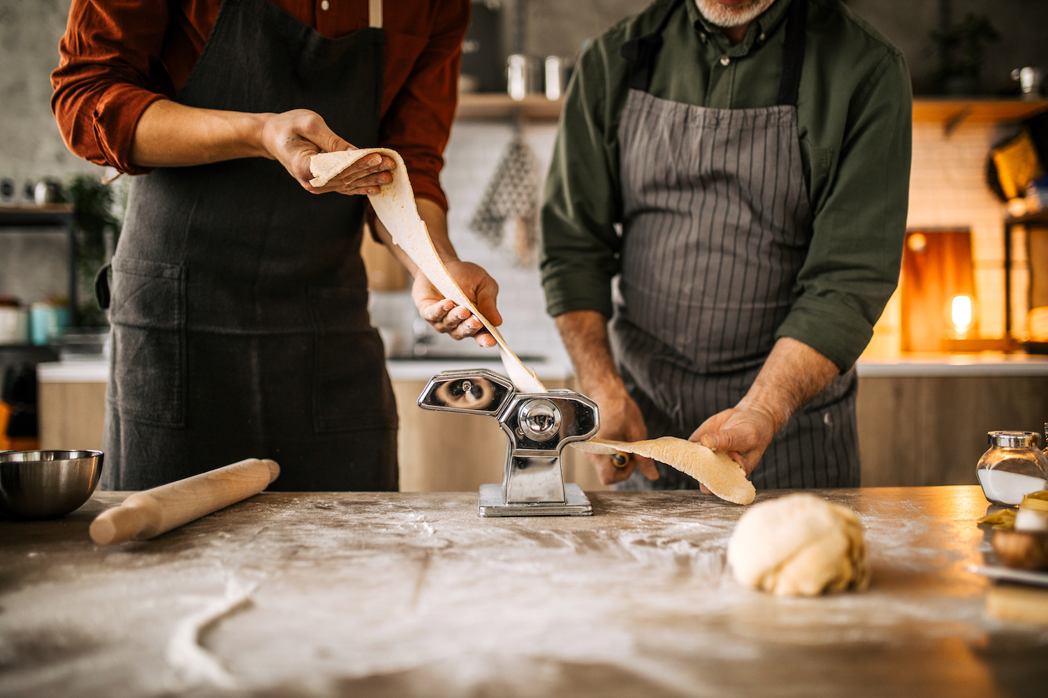 Senior father and young adult son making homemade pasta, rolling out the pasta dough using the manual pasta maker in domestic kitchen