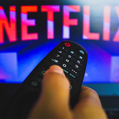 In this photo illustration, a hand holding a TV remote control in front of the Netflix logo on a TV screen.