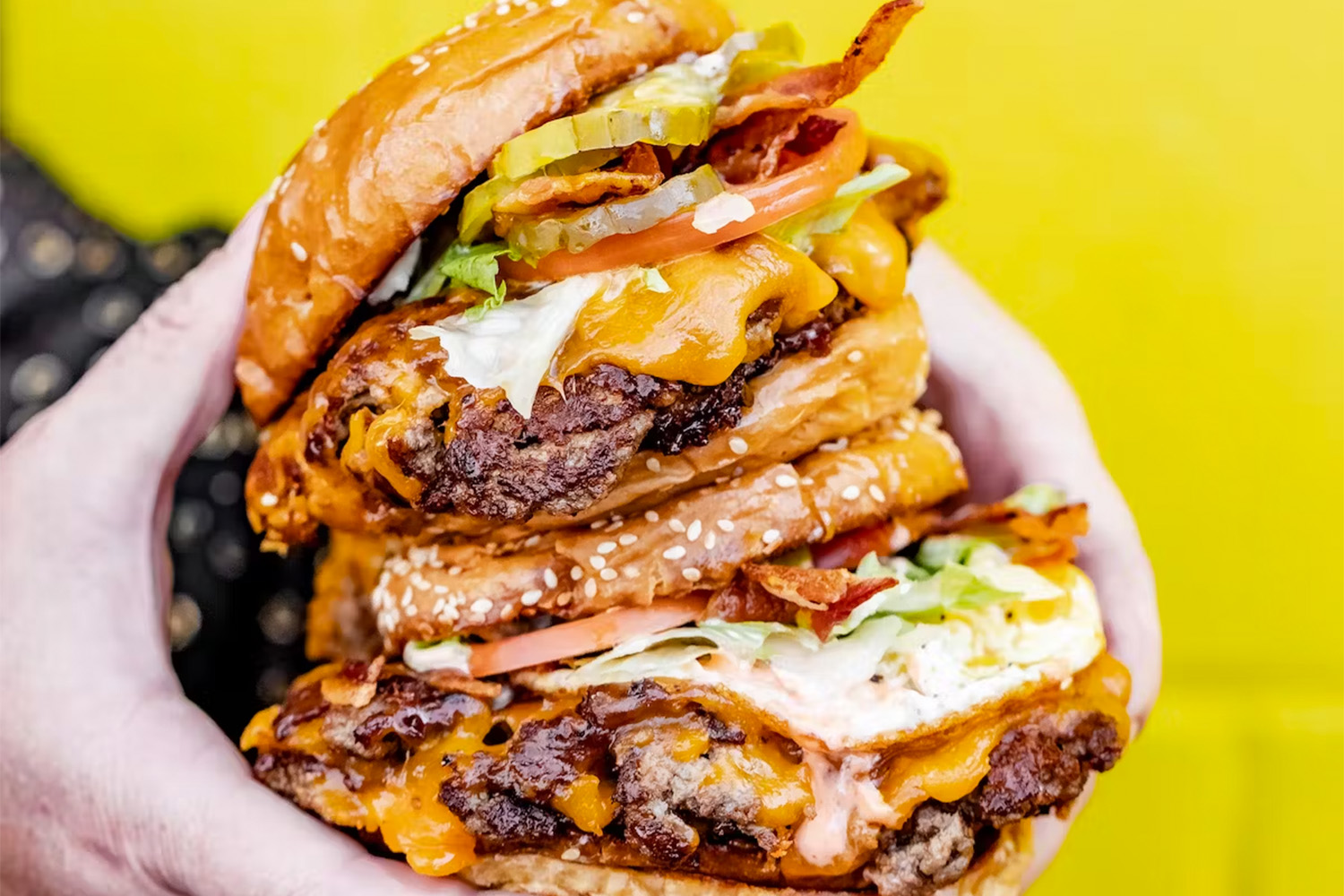 Two burgers being held in front of a yellow wall