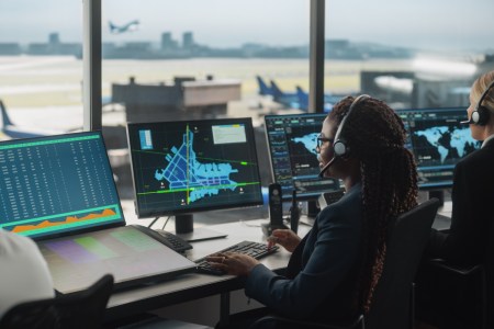 Female Air Traffic Controller with Headset Talk on a Call in Airport Tower. Office Room is Full of Desktop Computer Displays with Navigation Screens, Airplane Flight Radar Data for the Team