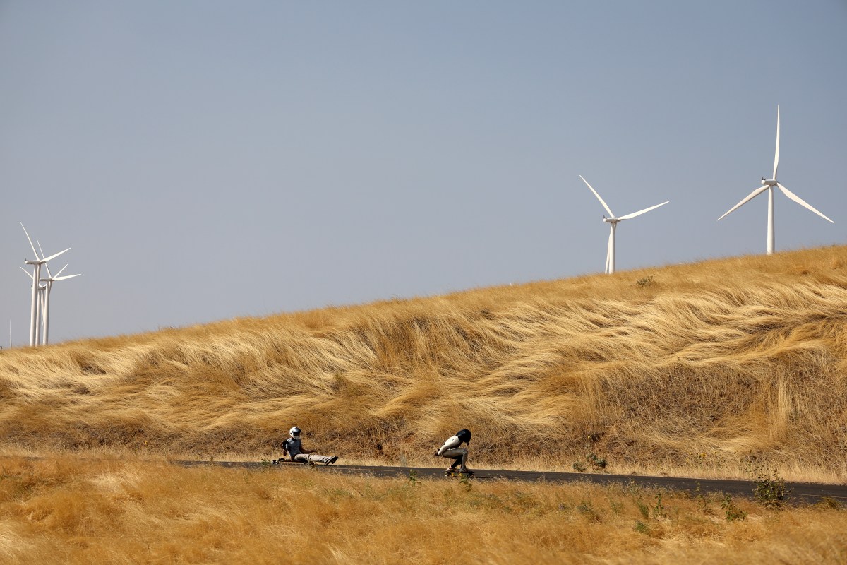 A view of a skater heading down a road at full speed, with yellow hills and windmills in the background.