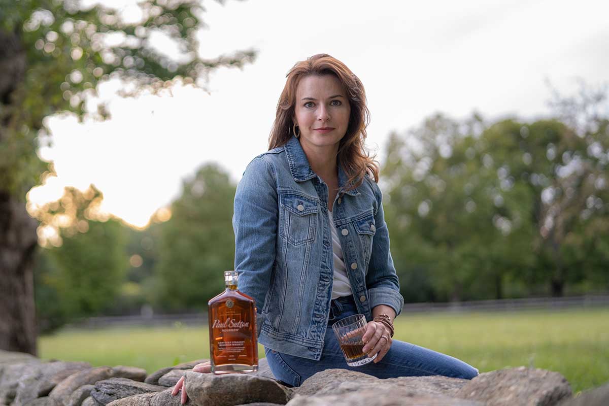 Paul Sutton CEO Myra Barginear sitting on a rock with a bottle and a glass of her bourbon