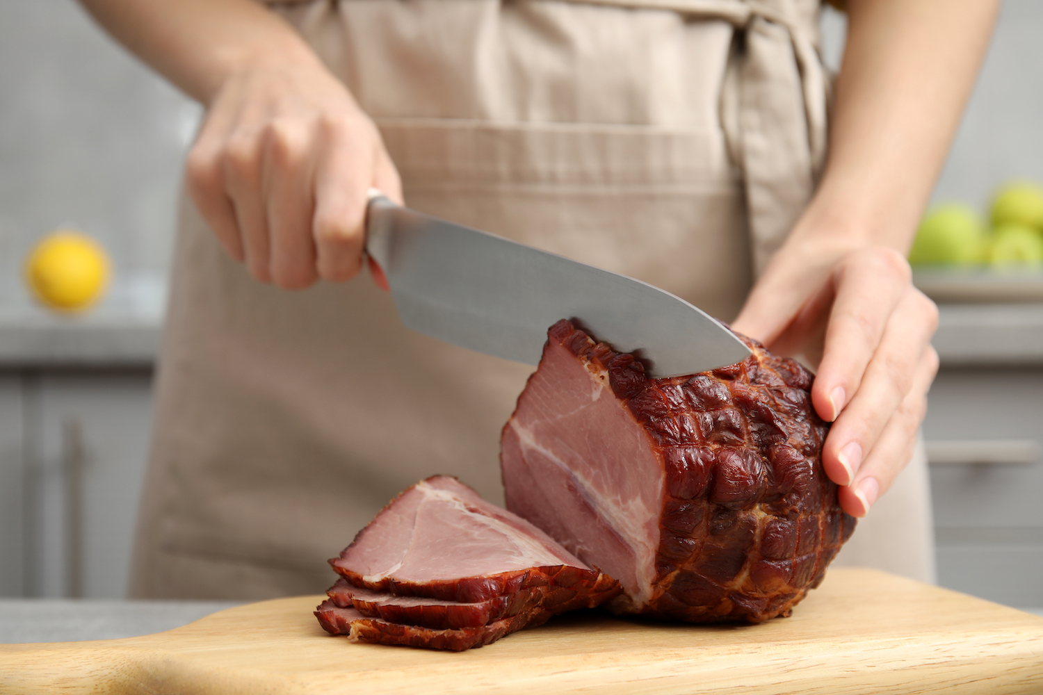 Woman cutting ham on wooden board at table indoors, closeup