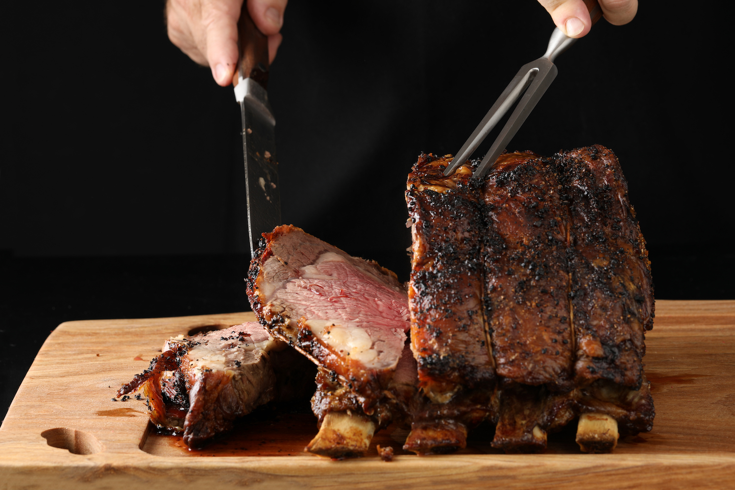 A close up horizontal photograph of a chefs hands carving a prime rib on a wooden carving board. Isolated on black