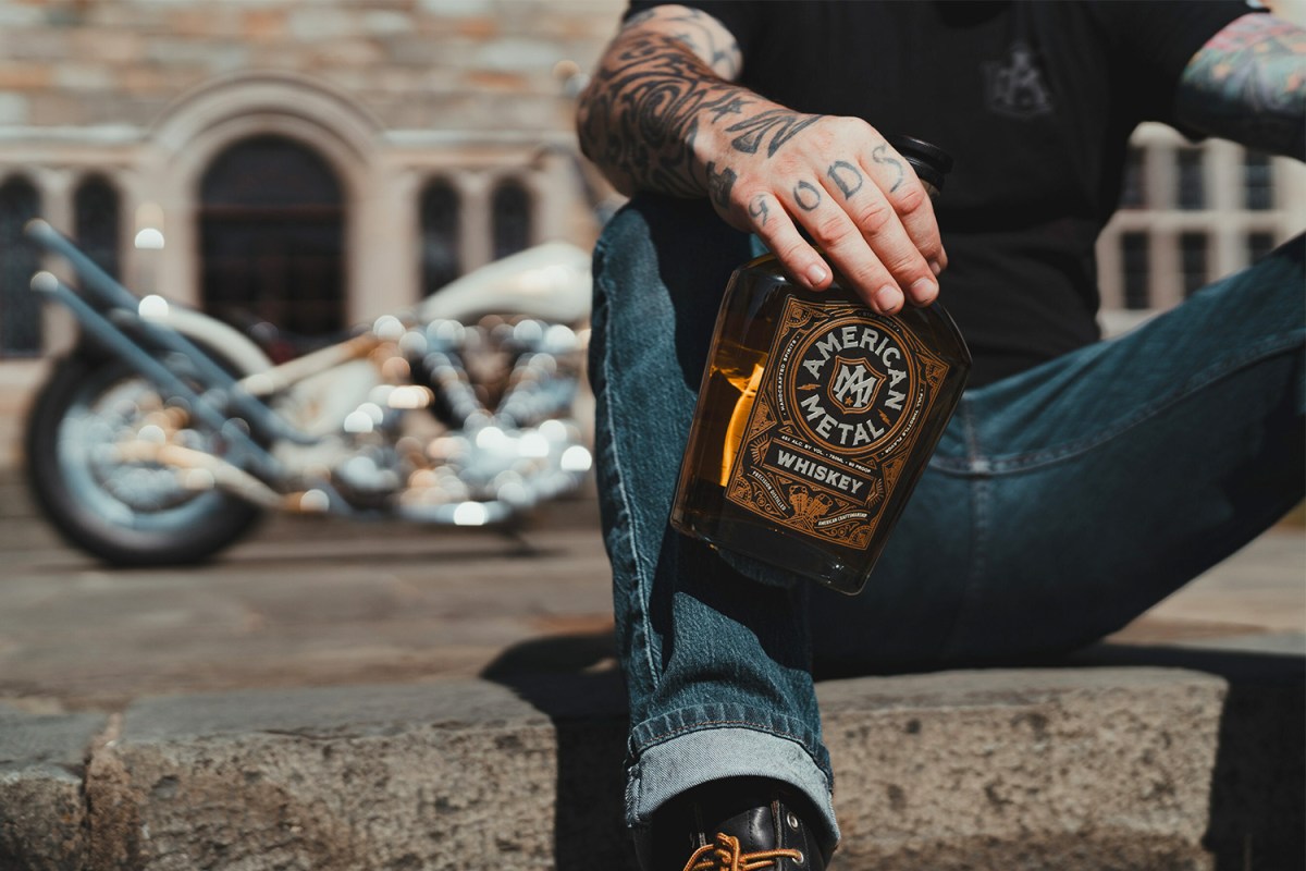 A man holding an American Metal Whiskey bottle near a motorcycle