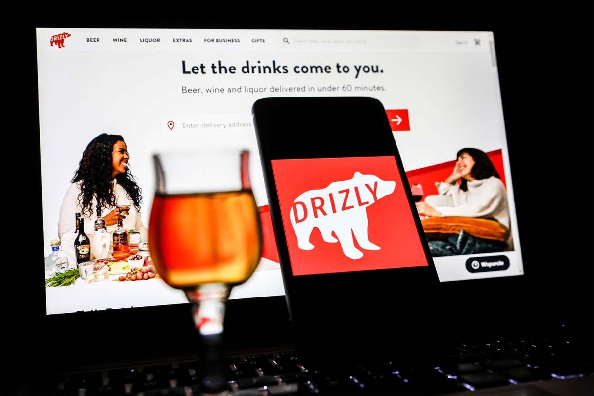 Drizly app logo is displayed on a mobile phone screen with Drizly website background. The booze delivery service is shutting down in March.