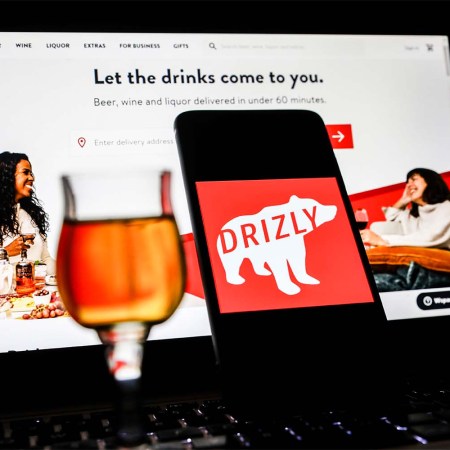 Drizly app logo is displayed on a mobile phone screen with Drizly website background. The booze delivery service is shutting down in March.