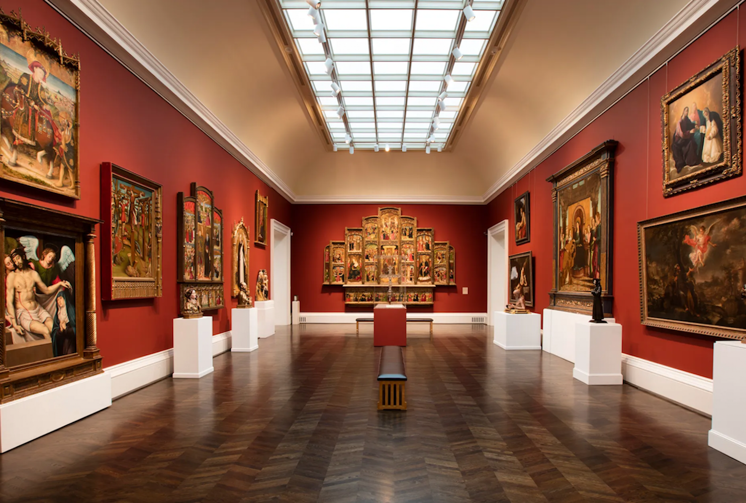 Hallway of a museum painted red with paintings hanging on the walls, podiums with statues on them and other tables in the cneter of the room