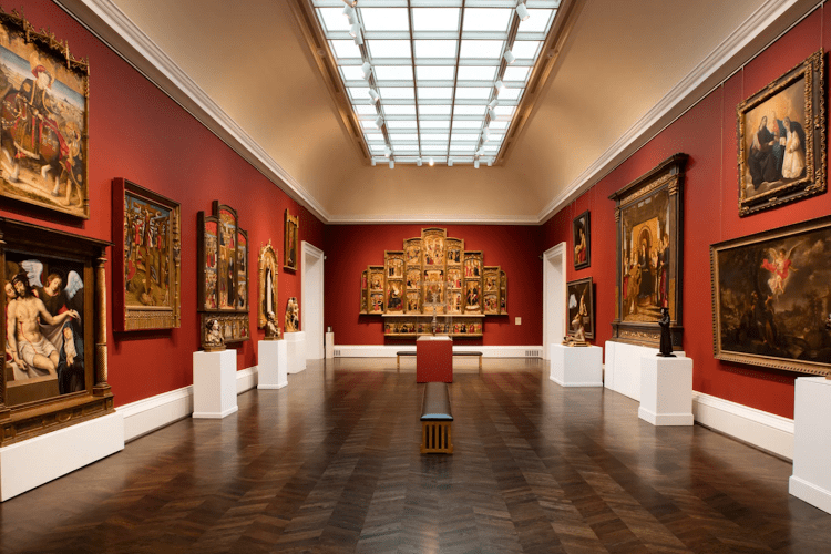 Hallway of a museum painted red with paintings hanging on the walls, podiums with statues on them and other tables in the cneter of the room