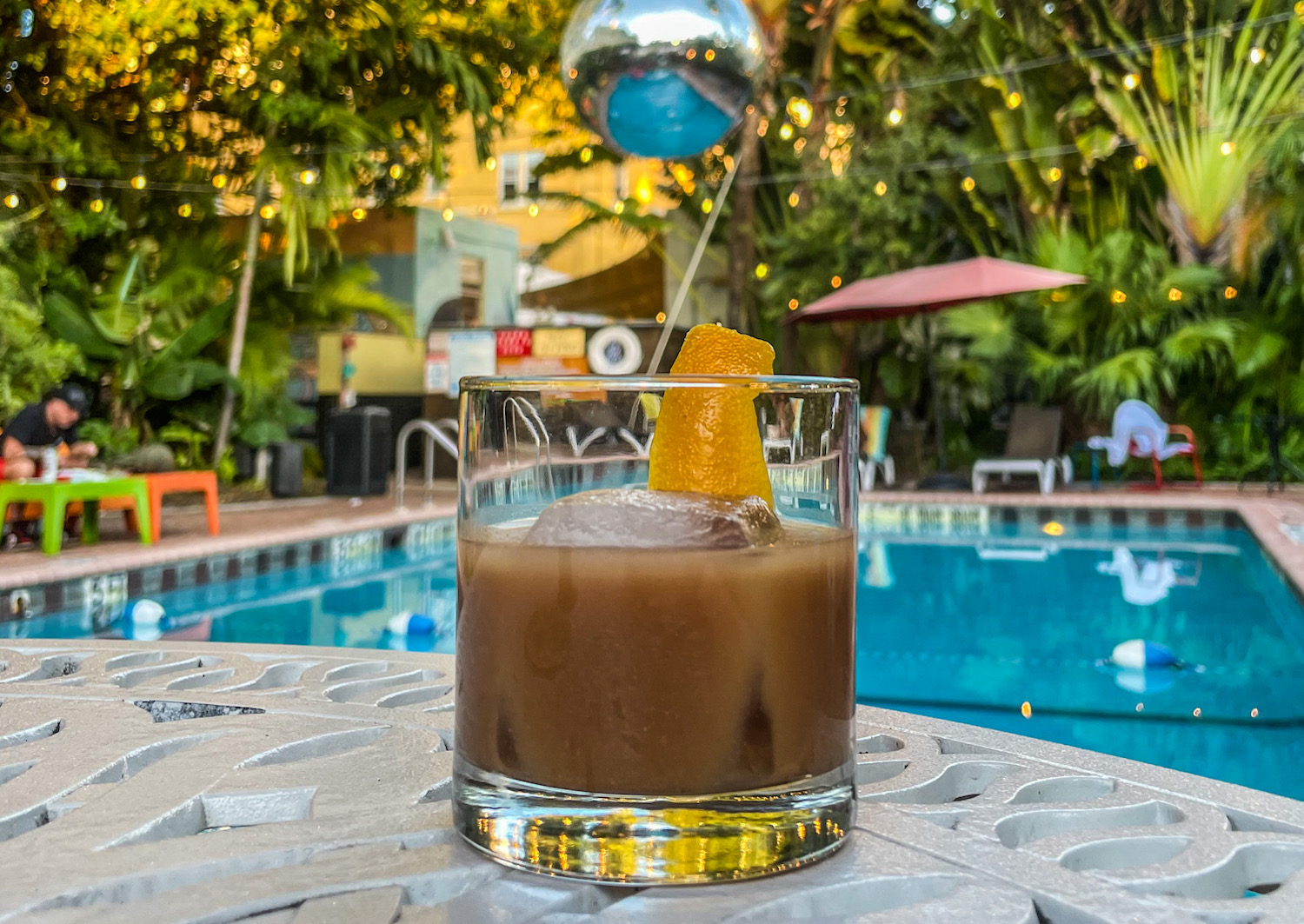 Brown liquid in a lowball glass with large sphere ice cube and orange peel twist in front of a pool 