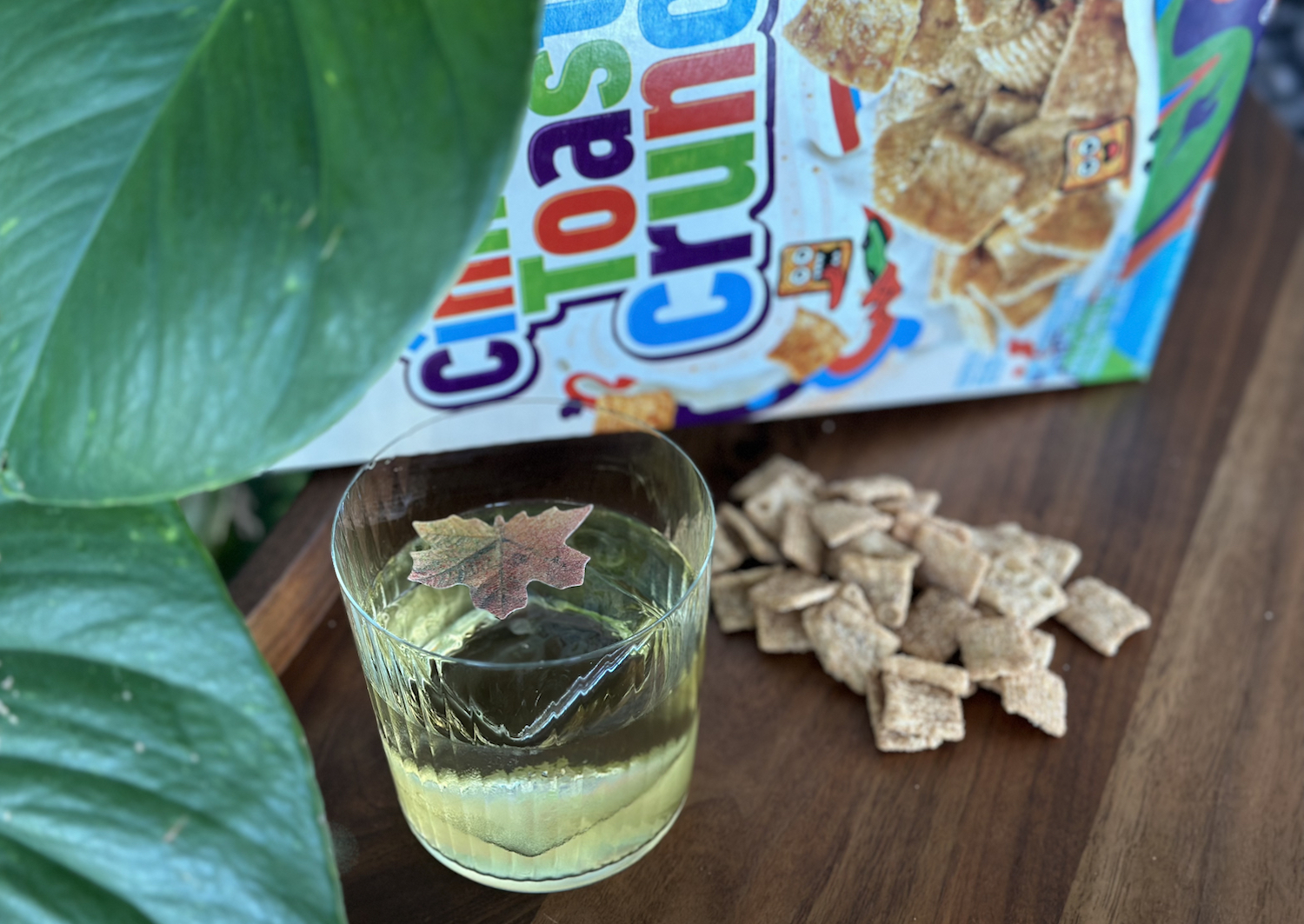 Lowball cocktail glass with clearish-green liquid and garnished with a brown leaf in front of a box of Cinnamon TOast Crunch and next to a plant. 