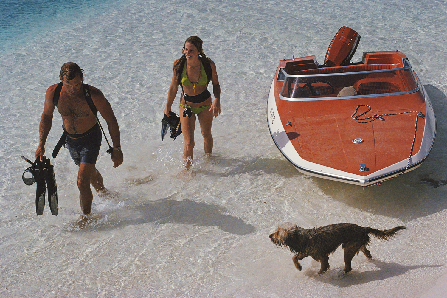 A man and woman return from snorkeling with their dog and boat on a tropical beach.