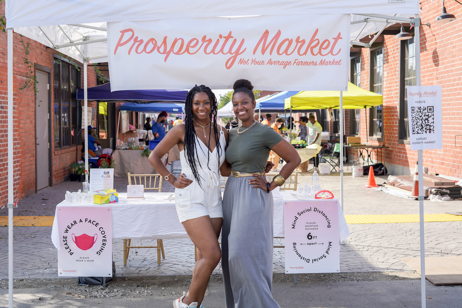 Carmen Dianne and Kara Still posing outside of their outdoor stand for their company Prosperity Market
