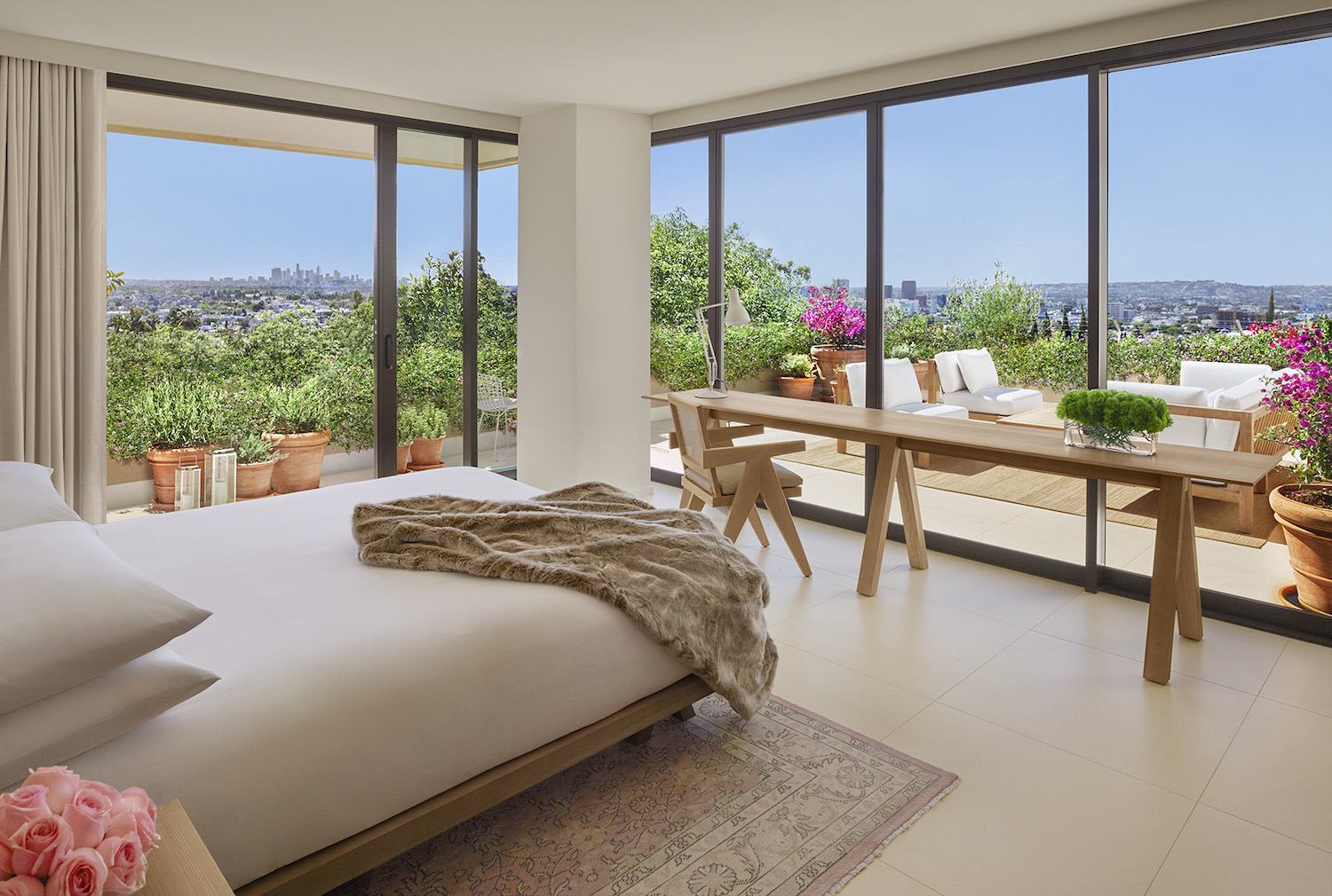 White bed in a room with floor-to-ceiling windows overlooking greenery and a terrace with a seating area that overlooks the city