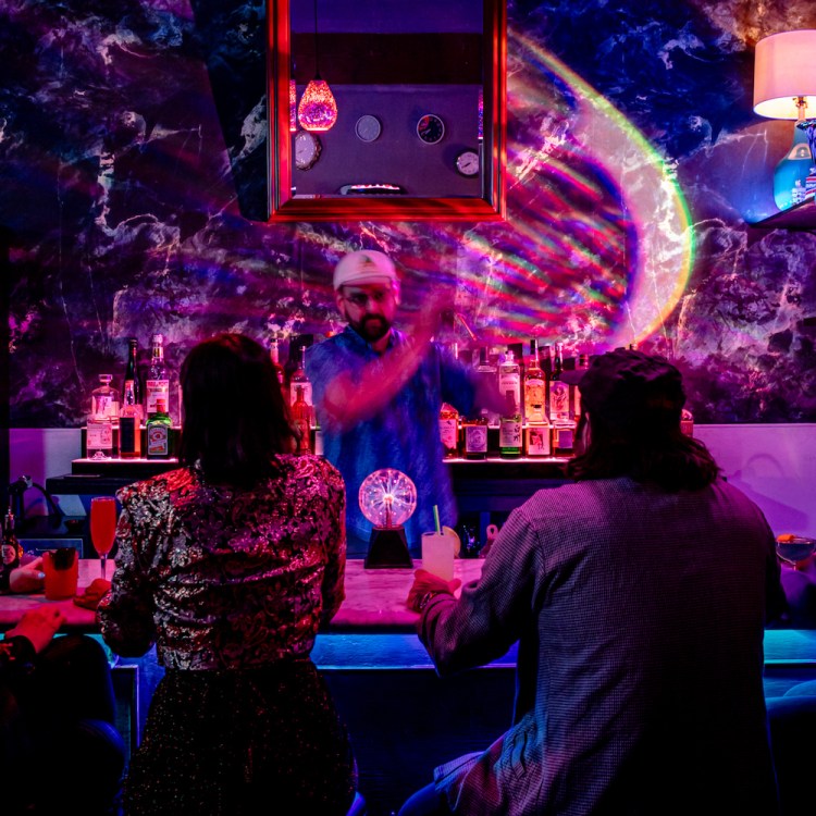 Bartender shaking a cocktail in front of people sitting at a bar in dim lighting with neon- colored disco ball lights