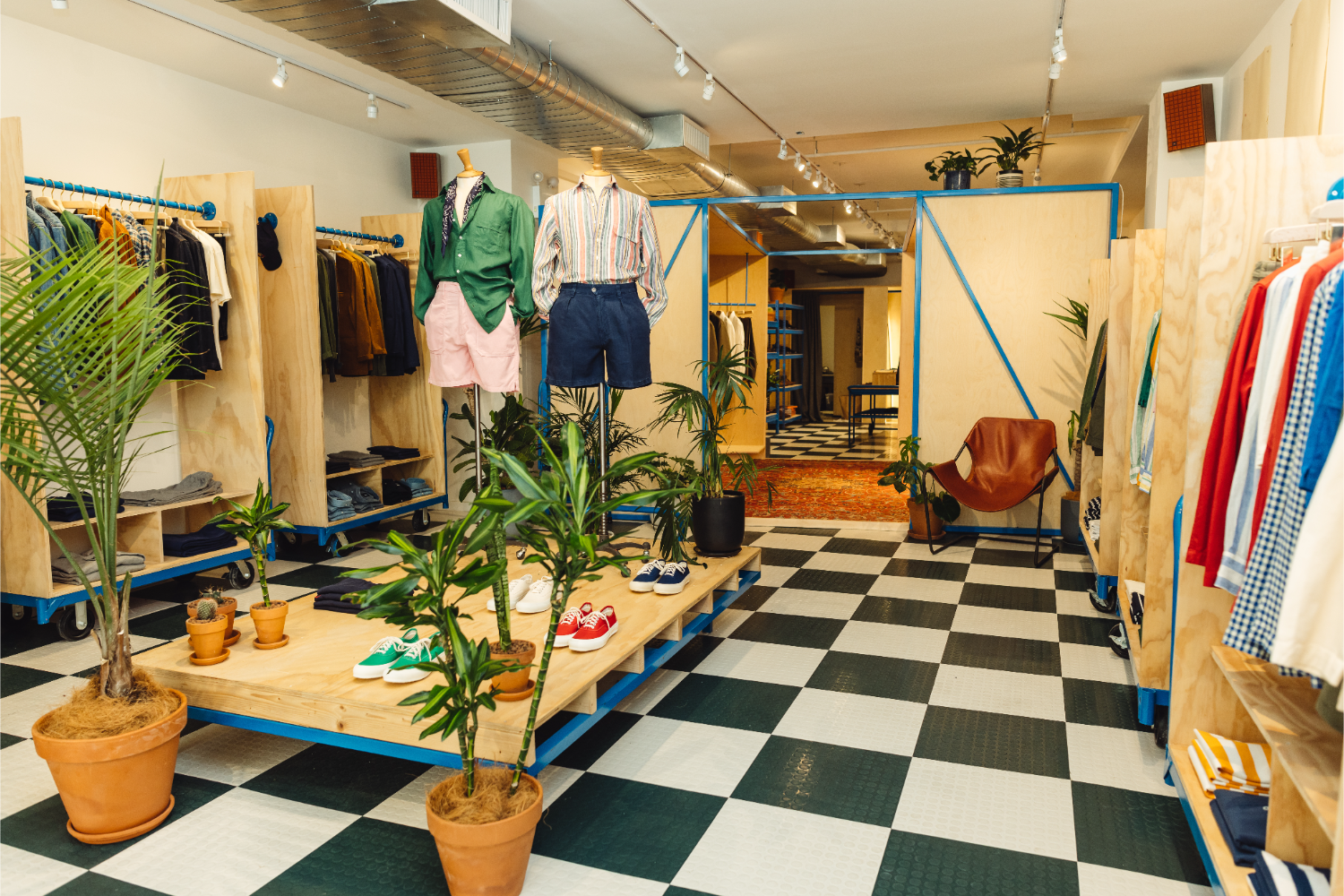 checkerboard floor, plants, mannequins with buttoned shirts and shorts, shoes on a wooden platform, clothes hanging 
