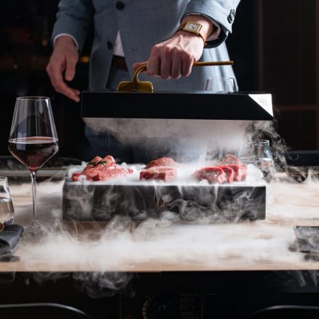 sliced red meat on a black box, a lot of smoke, man in gray suit holding black box top, wine glasses