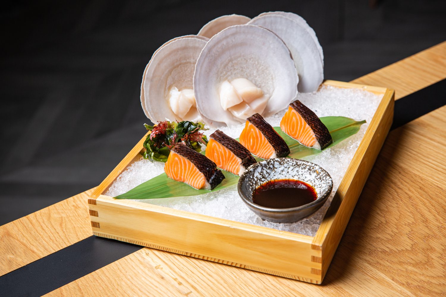 4 pieces of sushi on a leaf, 4 scallops, green garnish, sauce on the side, wooden box