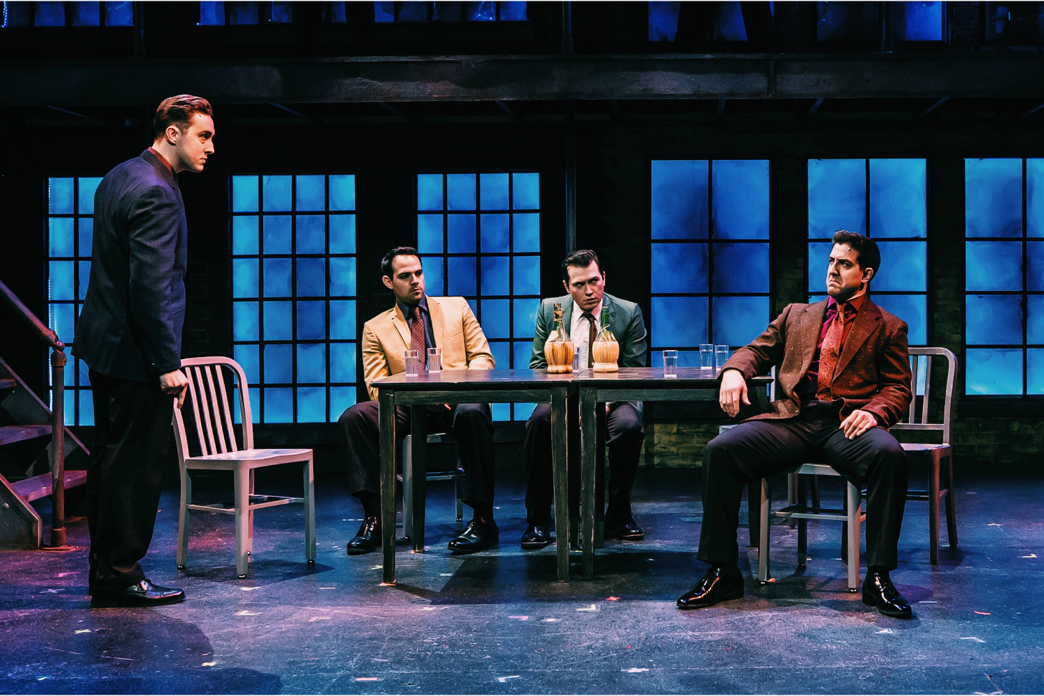 four male actors on stage in suits sitting in chairs, table  
