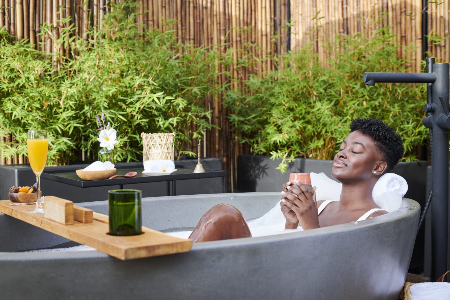 woman in bath tub outdoors with products on a stand, plants behind her 