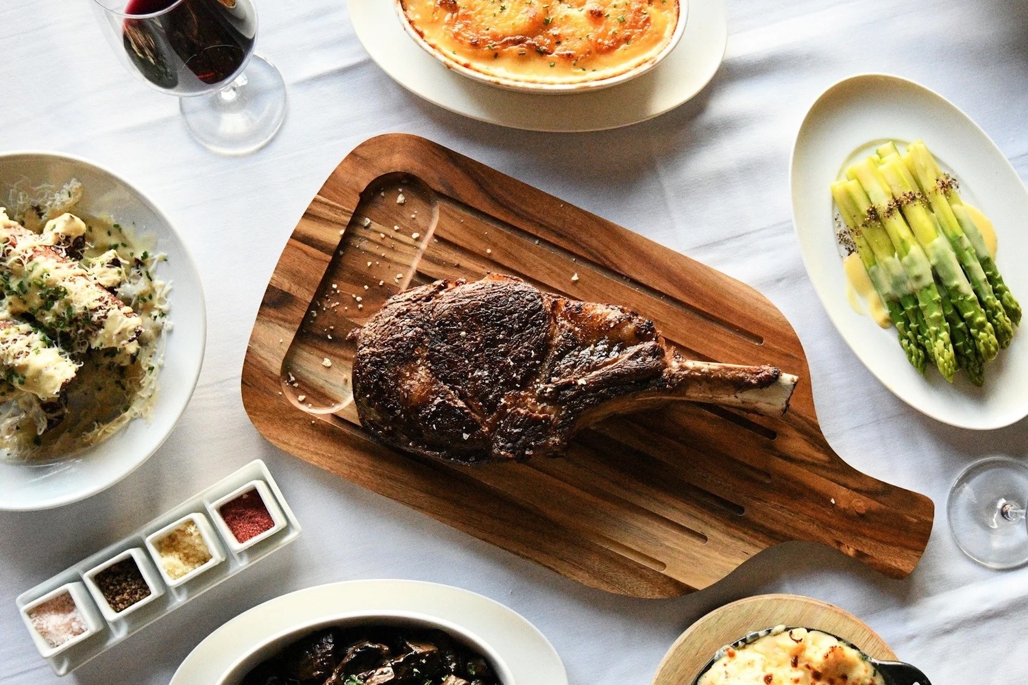 a steakhouse spread on a white tablecloth including steak, asparagus, manicotti and red wine
