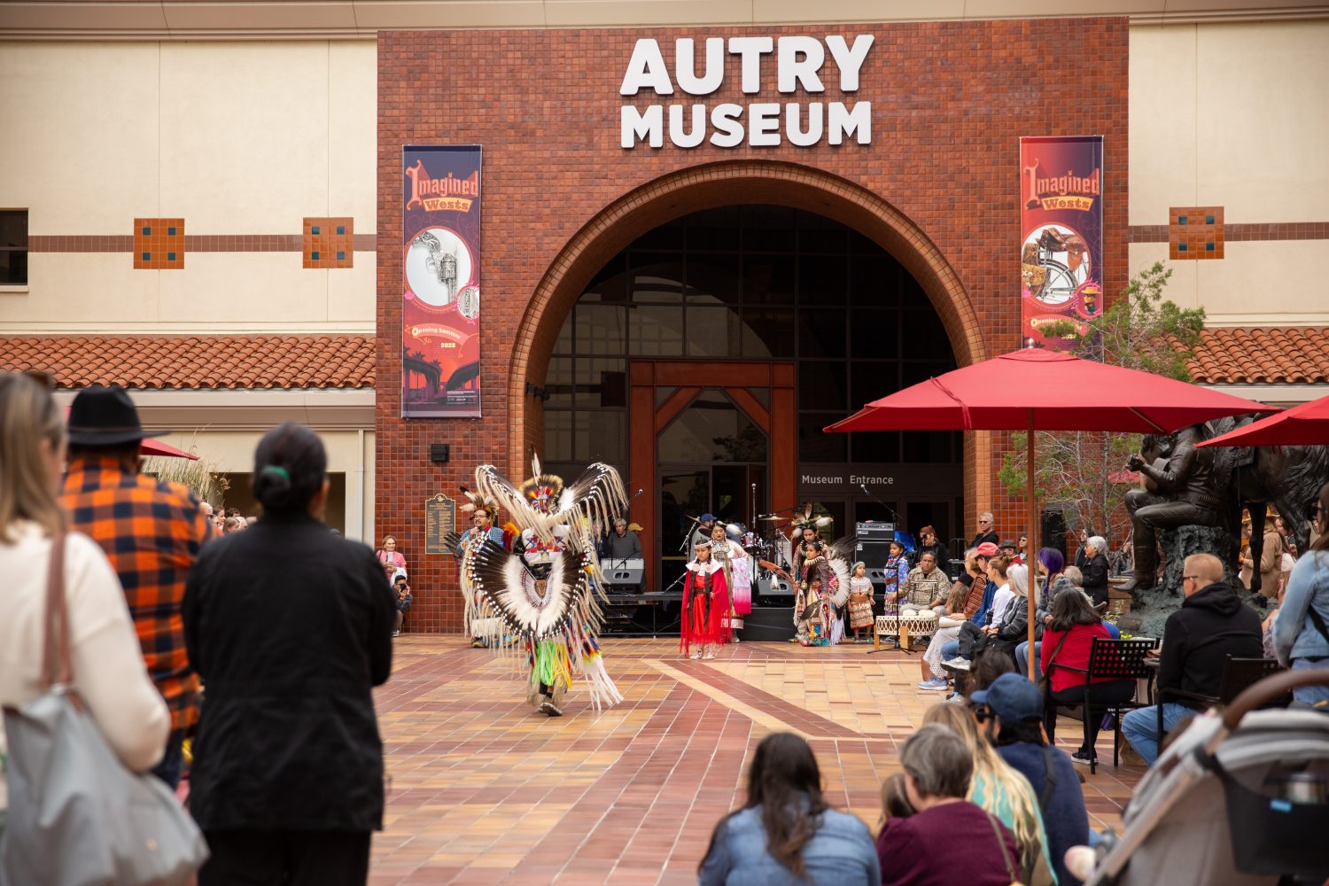 people sitting watching person in traditional native american dress perform outside museum entrance