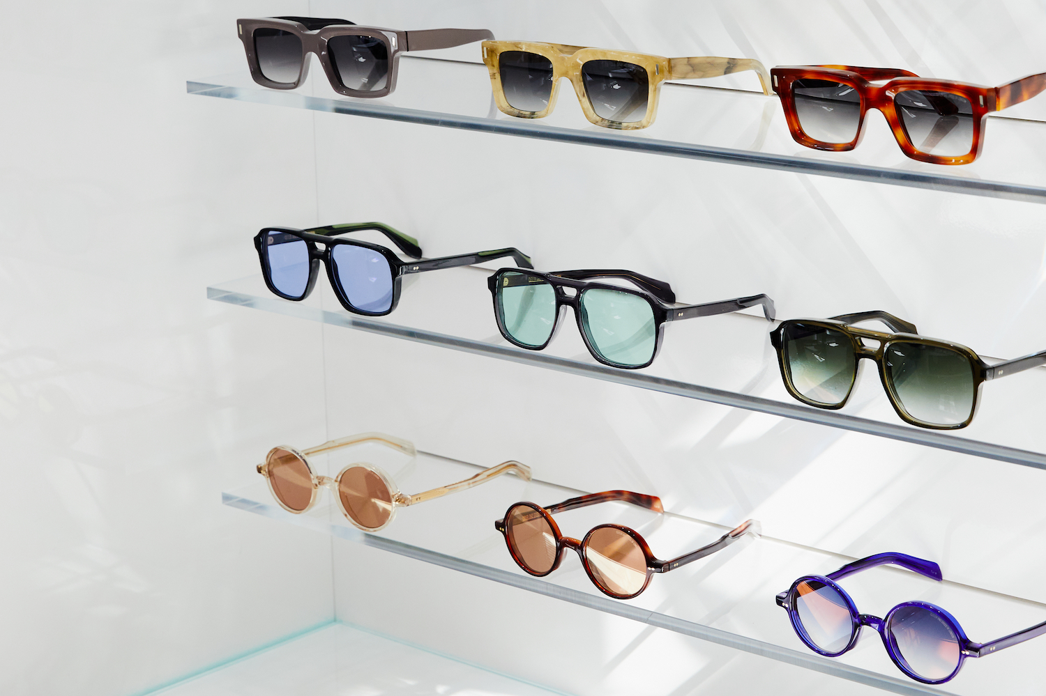 three rows of sunglasses on glass shelves in front of a white wall