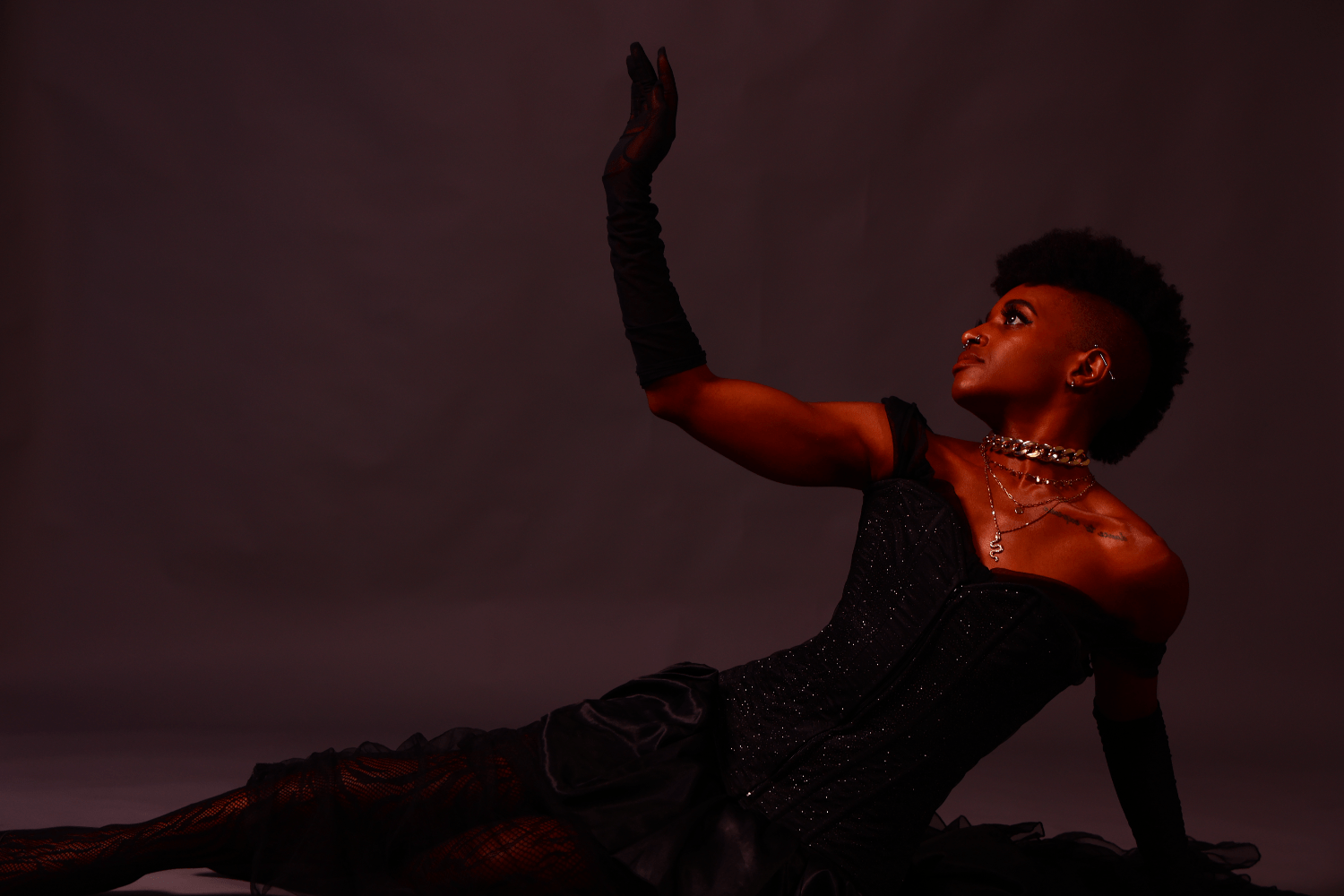 woman with piercings, silver jewelry, tattoo on collarbone and  frohawk, in a black shimmery dress laying down sideways while looking up, arm out