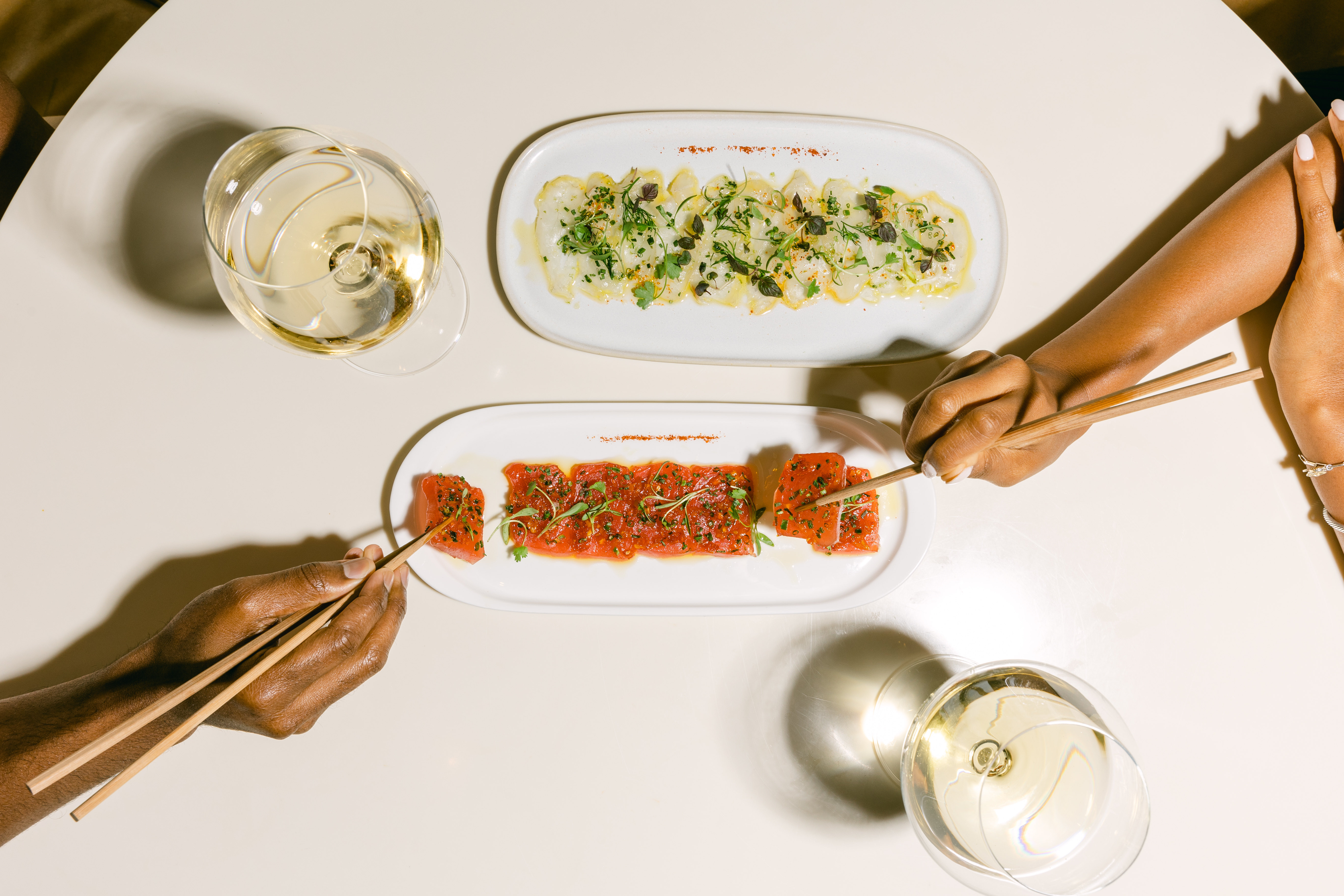 two hands holding chopsticks, sashimi on plate, wine glasses