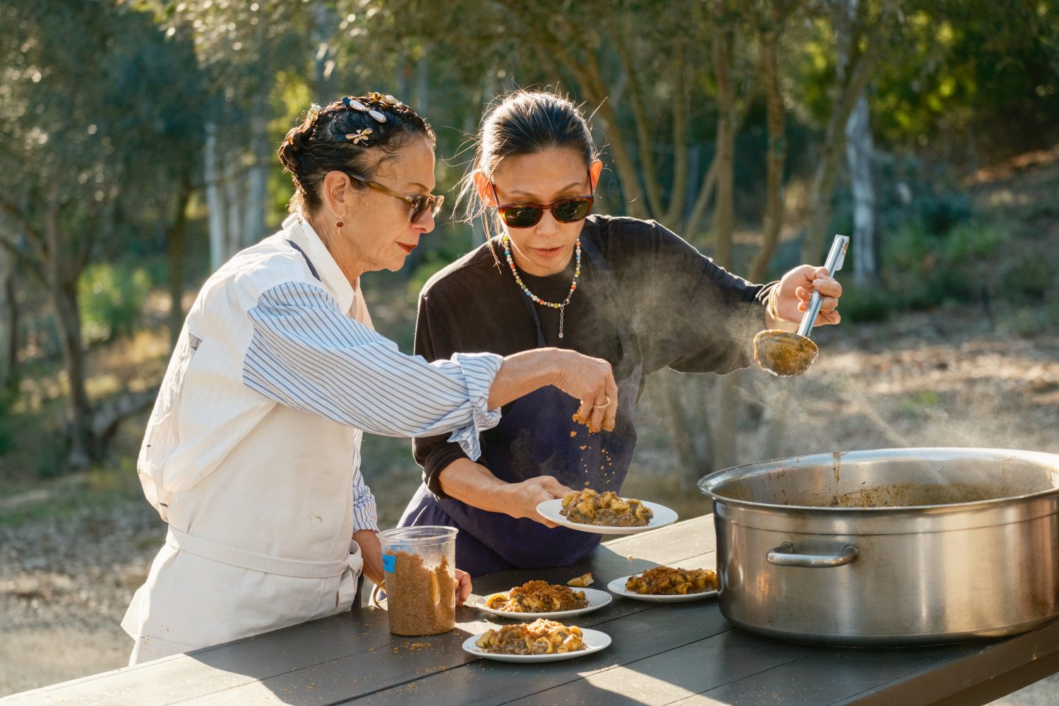 two women with sunglasses serving food onto plates from a steaming pot