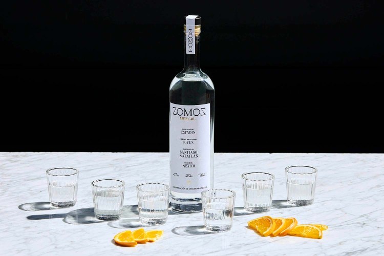 A bottle of Zomoz mezcal near orange slices and six shot glasses