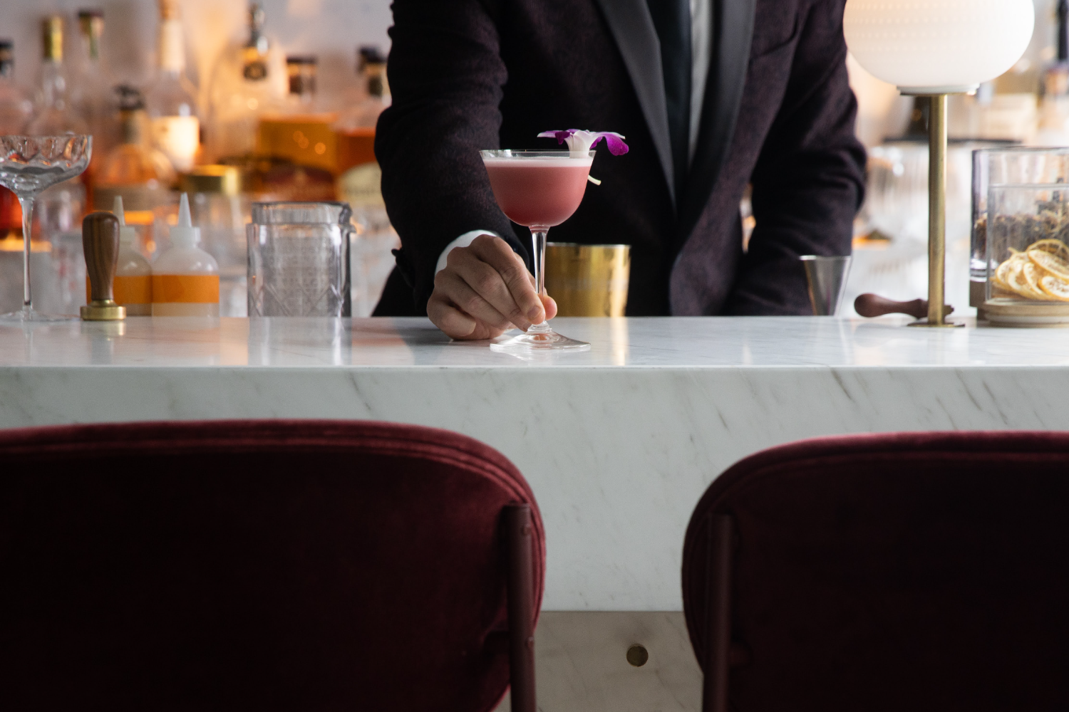 man in suit holding pink cocktail with flower in it, burgundy chairs, white marble bar