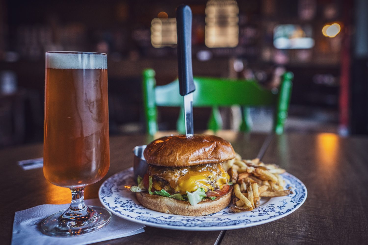 burger with knife going through middle, side of fries, beer on the side, green chair in background