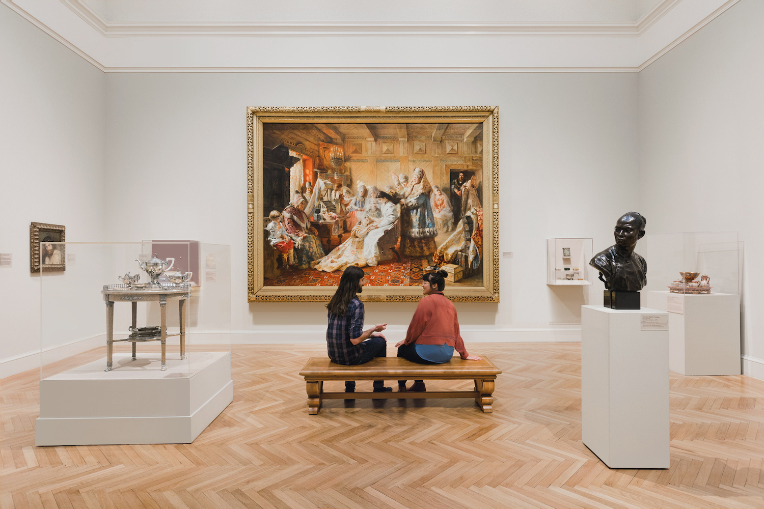two people sitting on a bench in a museum talking and admiring a gold-framed painting