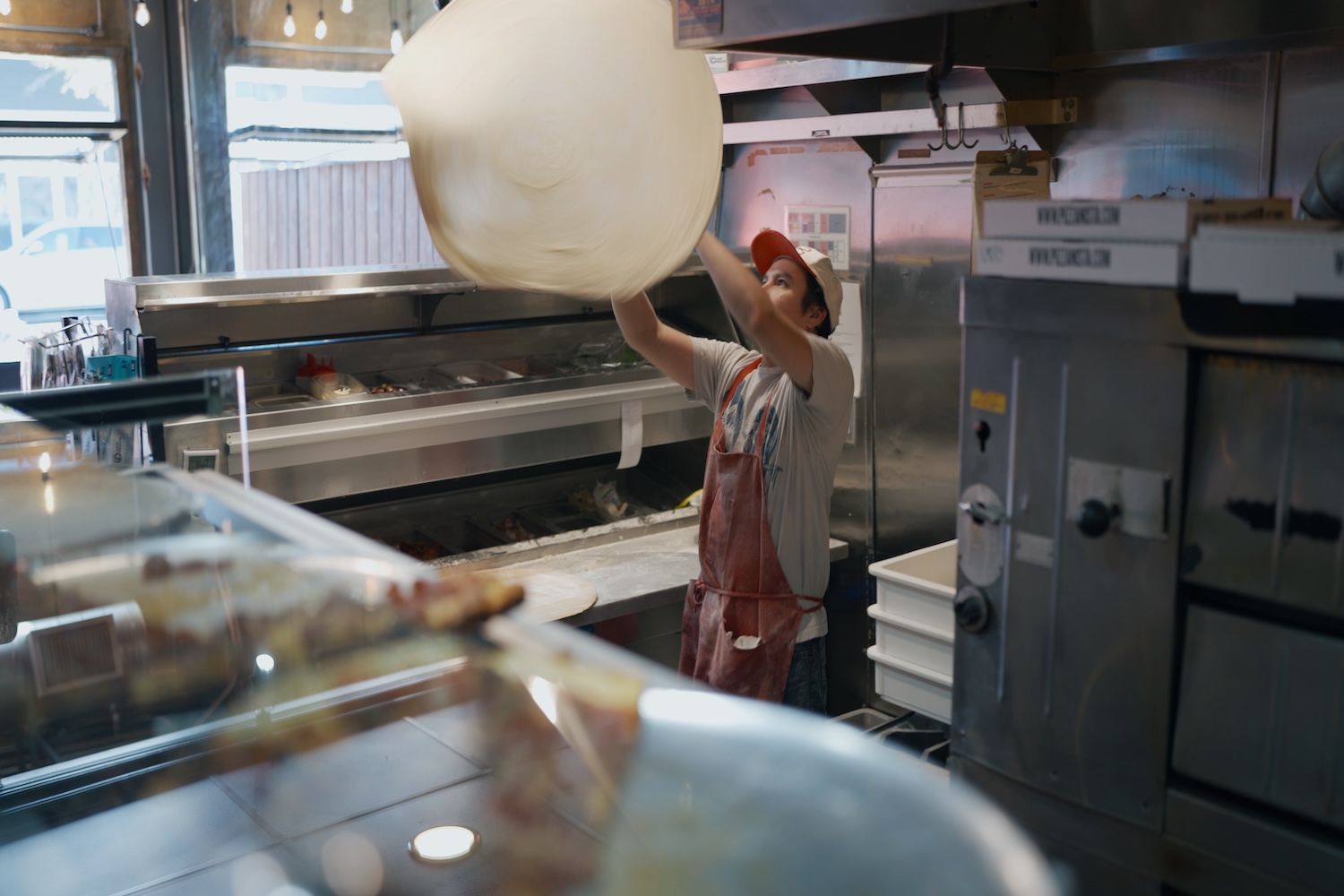 man in hat and apron throwing dough in air, in a kitchen