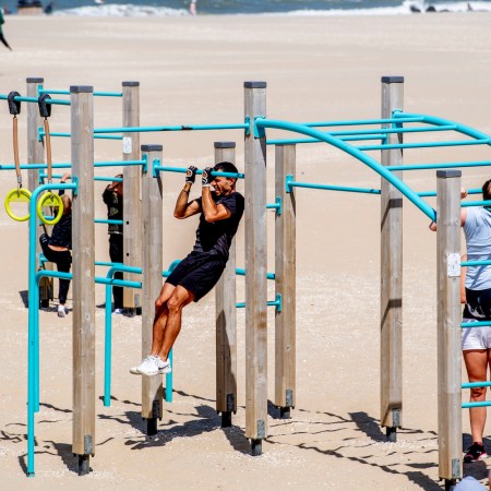 Two women, two kids and one man climbing and doing pull-ups on a jungle gym with blue bars on the sand