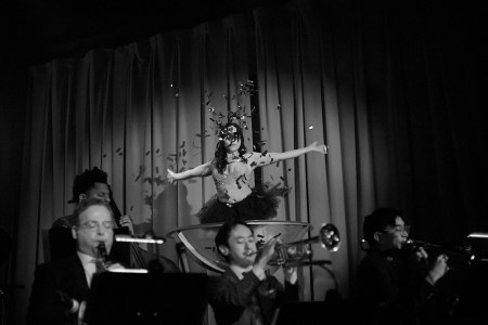 woman in a giant glass wearing a tutu throwing confetti, men playing instruments in suits