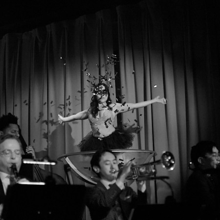 woman in a giant glass wearing a tutu throwing confetti, men playing instruments in suits