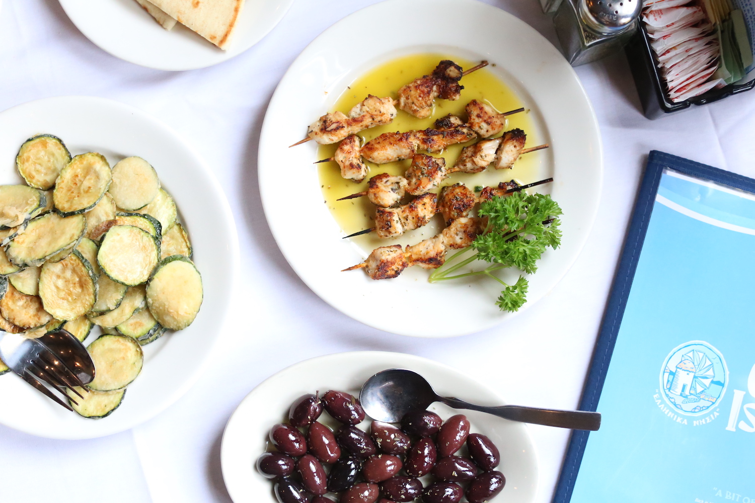 chicken kabobs, fried zucchini and kalamata olives on white plates sitting on a white tablecloth