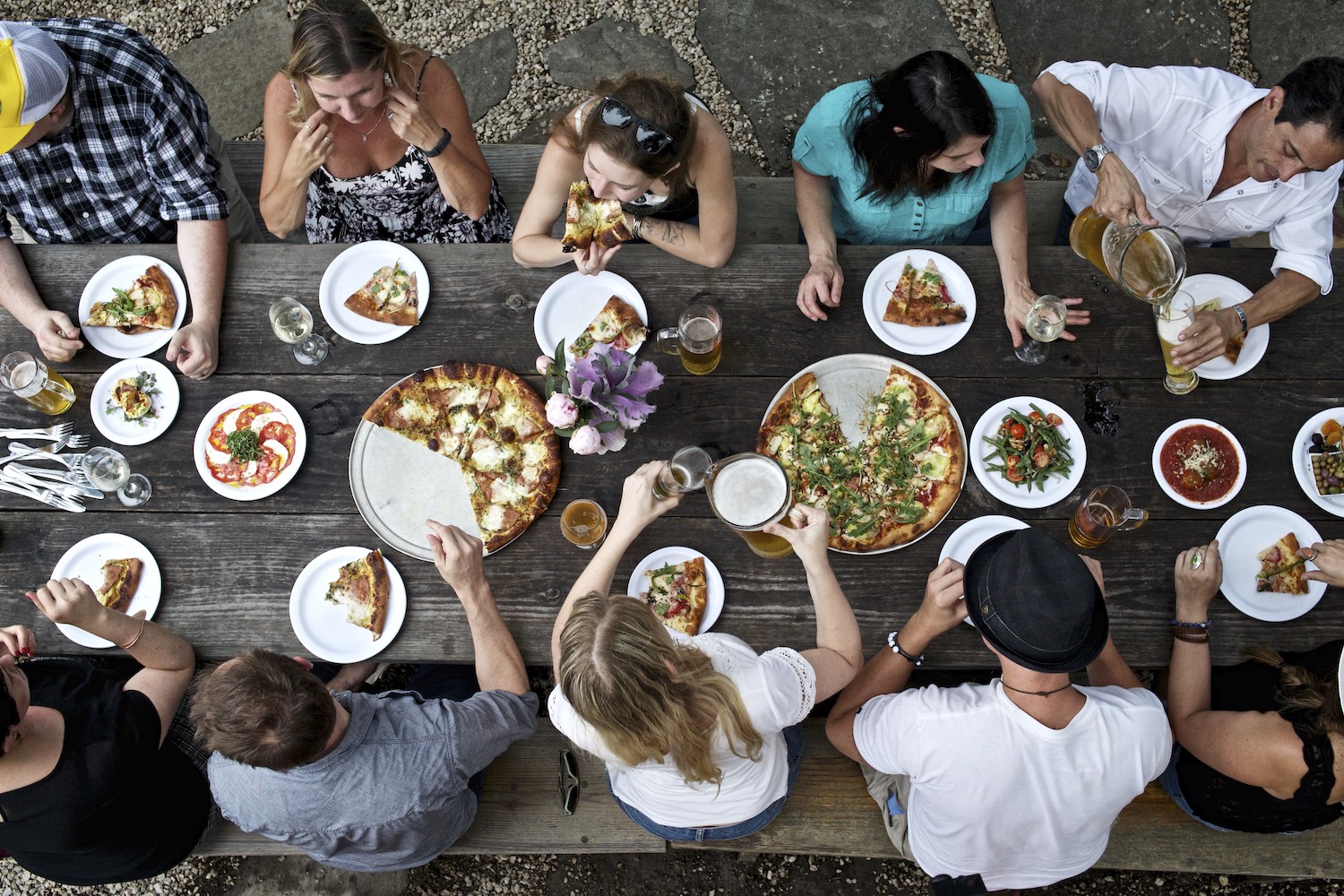 10 people sitting at a long wooden table eating pizza and drinking beer