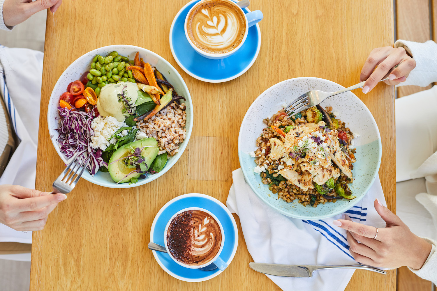 two people sitting at a wooden table eating bowls of food and drinking lattes out of blue ceramic mugs