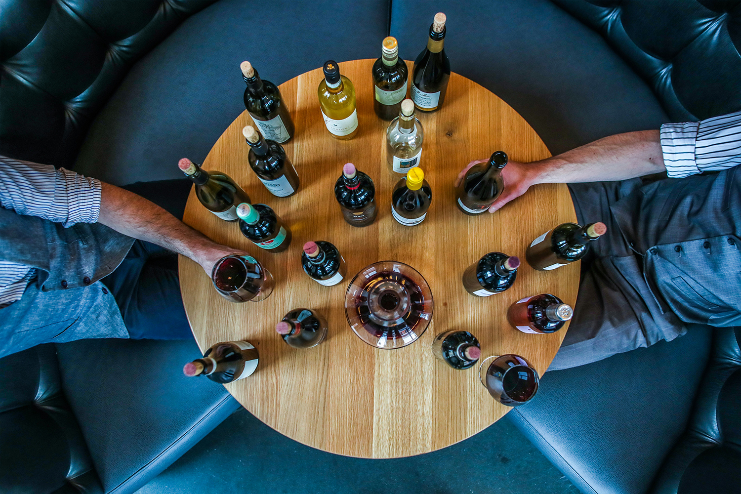 Two men sitting across from each other at a small round wooden table with a bunch of bottles and glasses of wine. Today we recommend 14 bottles of chilled red wine to drink this summer.